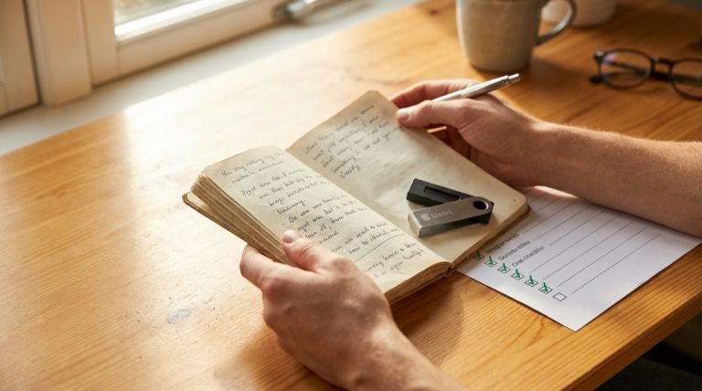 Person verifying handwritten seed phrase words against a hardware wallet with a checklist on a wooden desk
